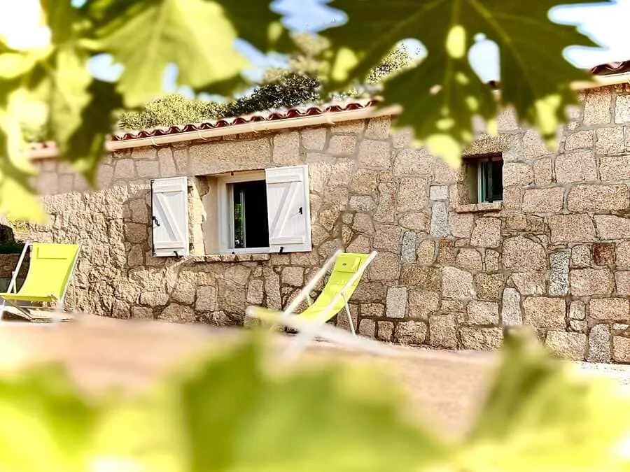 Façade en pierre de la maison de vacances en Corse, avec deux transats jaunes disposés à l'extérieur sous des feuillages.