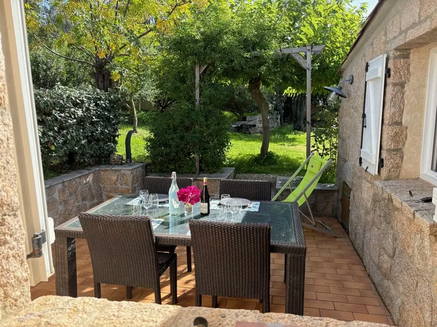 Terrasse extérieure avec table en rotin, verres et bouteille de vin, entourée de verdure, maison de vacances en Corse.